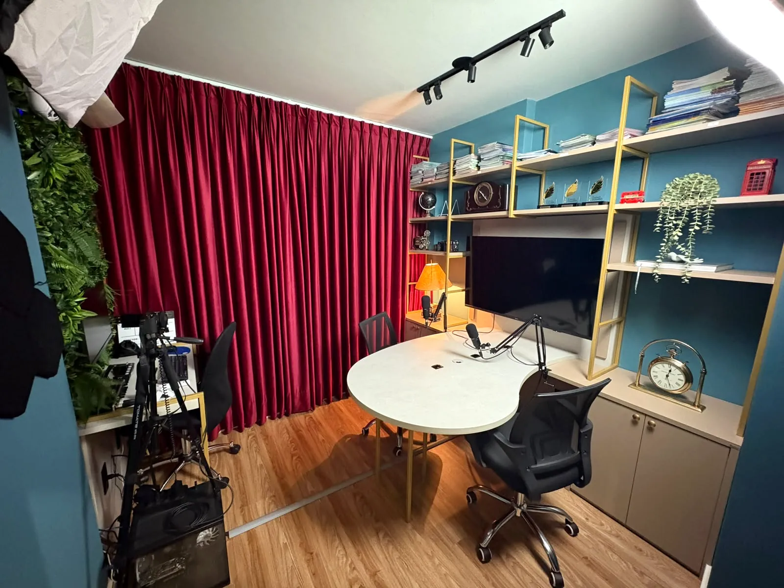 Estúdio de podcast com mesa orgânica, microfones, cortina de veludo bordô e estante com telão